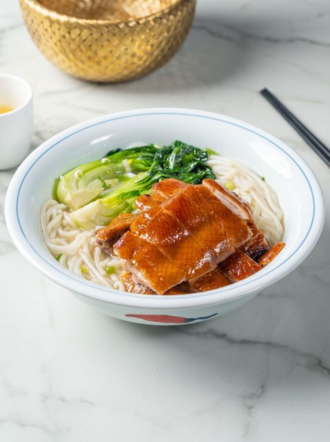 Bowl of noodle soup with roasted pork and greens on a marble table