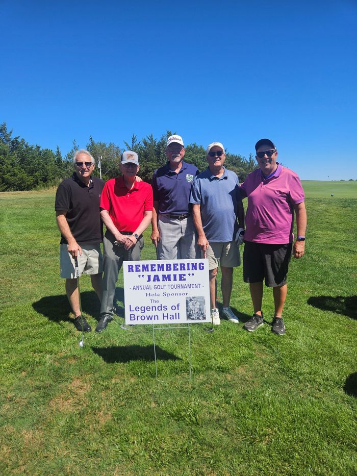 A group of men are standing in a grassy field holding a sign for the remembering jamie golf tournament