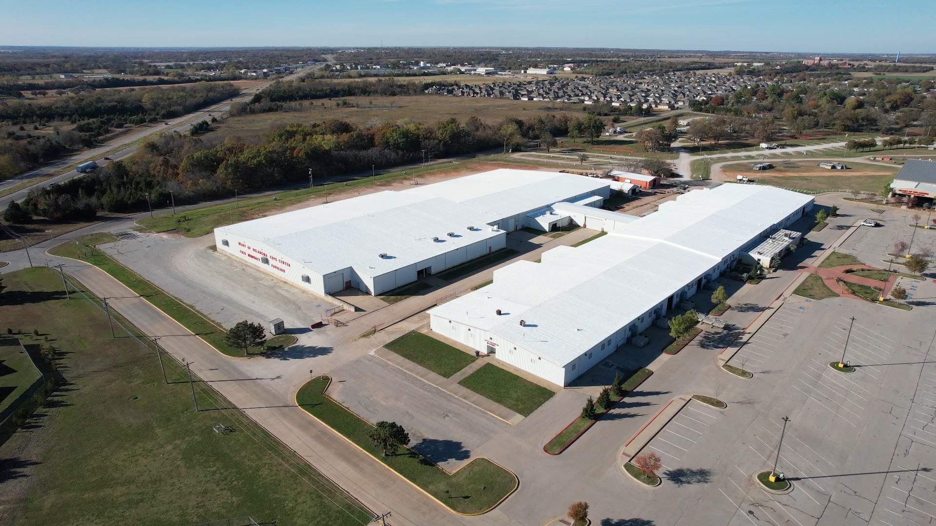 Aerial view of two large white-roofed buildings with surrounding parking and greenery under a clear blue sky.