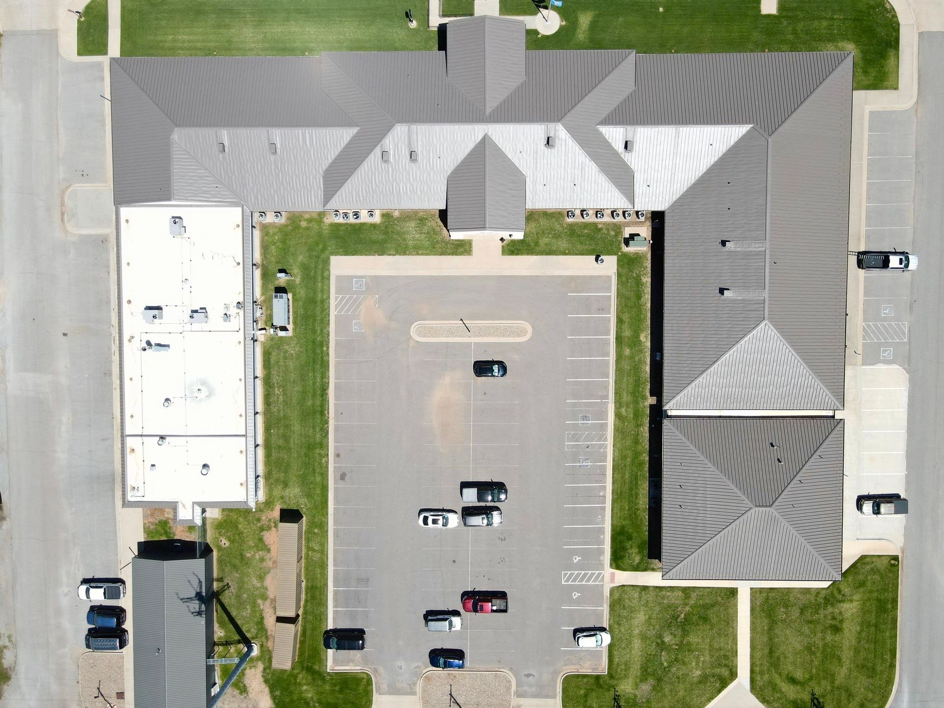 Aerial view of a large building with a gray roof, parking lot with cars, and surrounding grass and street.