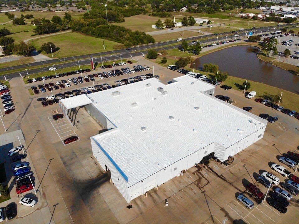 Aerial view of a car dealership with white building, cars in parking lot, highway, and a pond.