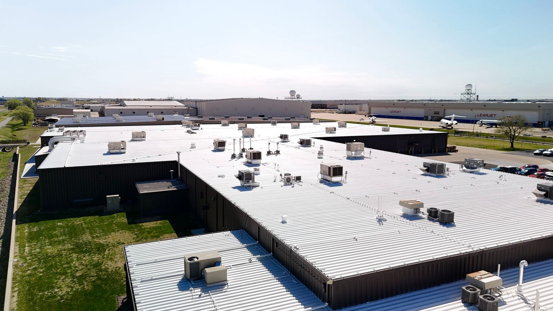 An aerial view of a flat, light-colored industrial warehouse roof featuring numerous HVAC units on a sunny day.