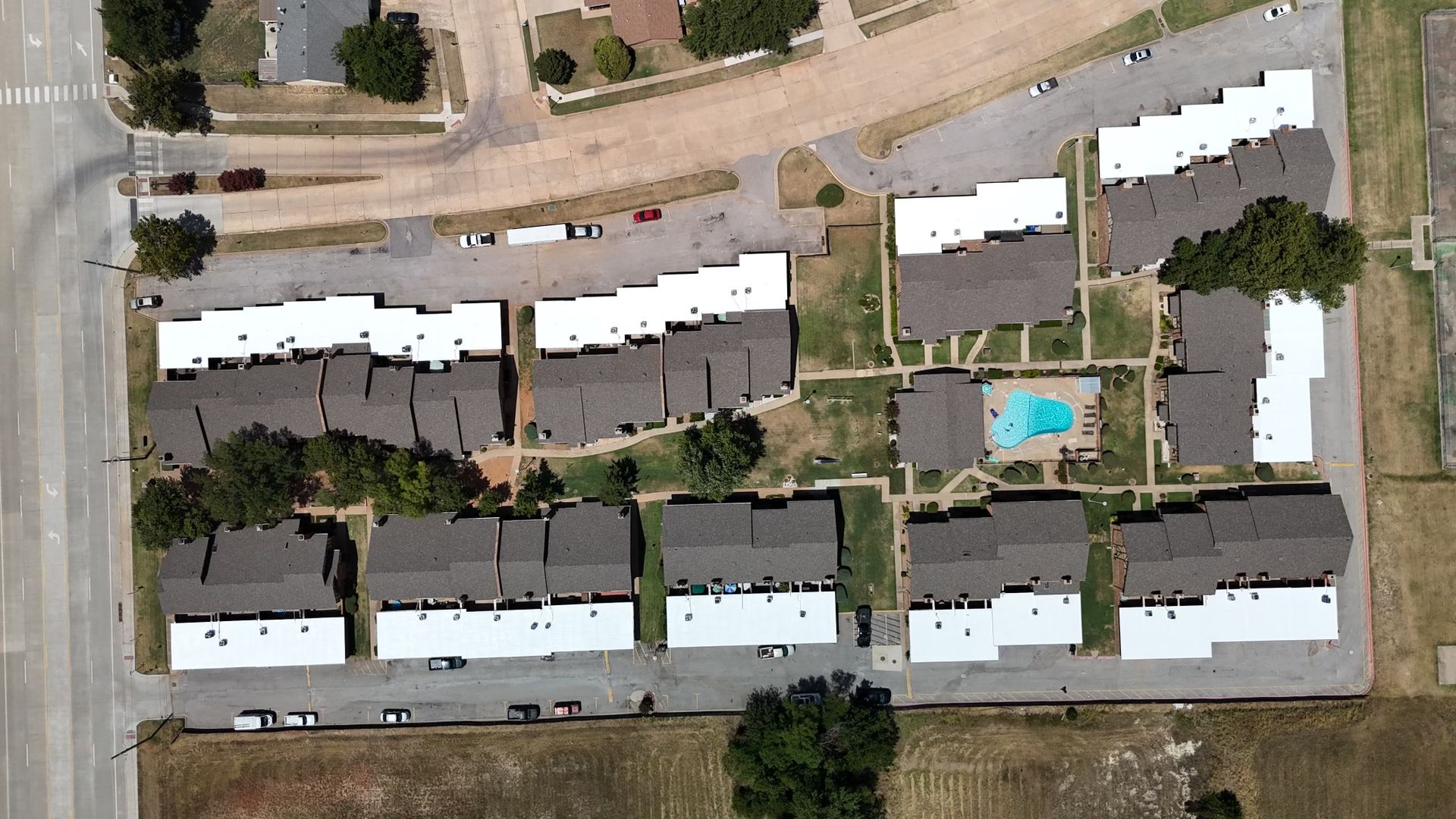 An aerial view of an apartment complex with multiple buildings, paved parking lots, and a small central swimming pool.