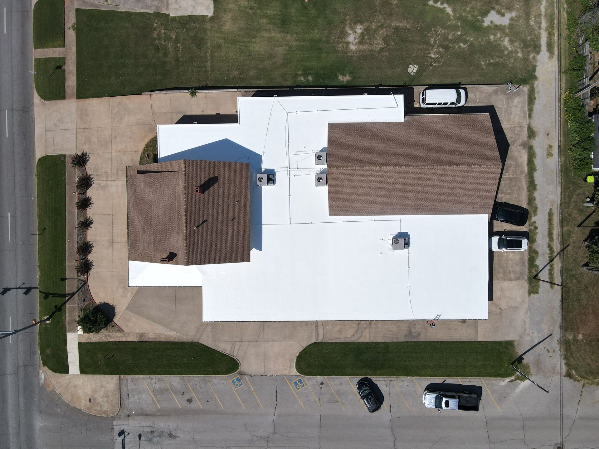 Aerial view of a building with a white flat roof and two brown pitched-roof sections, situated on a paved lot.