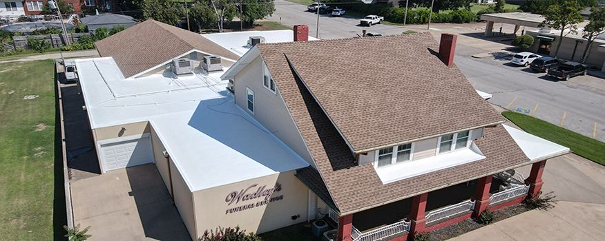 Aerial view of a brown-roofed building with white walls. A driveway and cars are to the right.