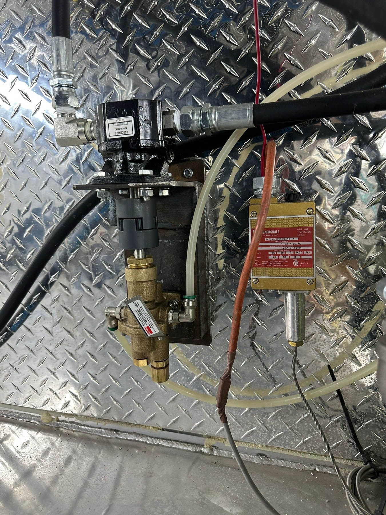 A brass solenoid valve and a hydraulic component mounted on a diamond-plate metal wall with attached tubing and wires.