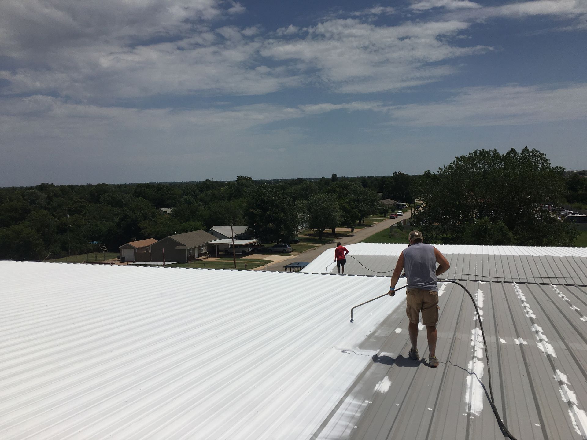 Two workers coat a corrugated metal roof with white sealant on a sunny day.