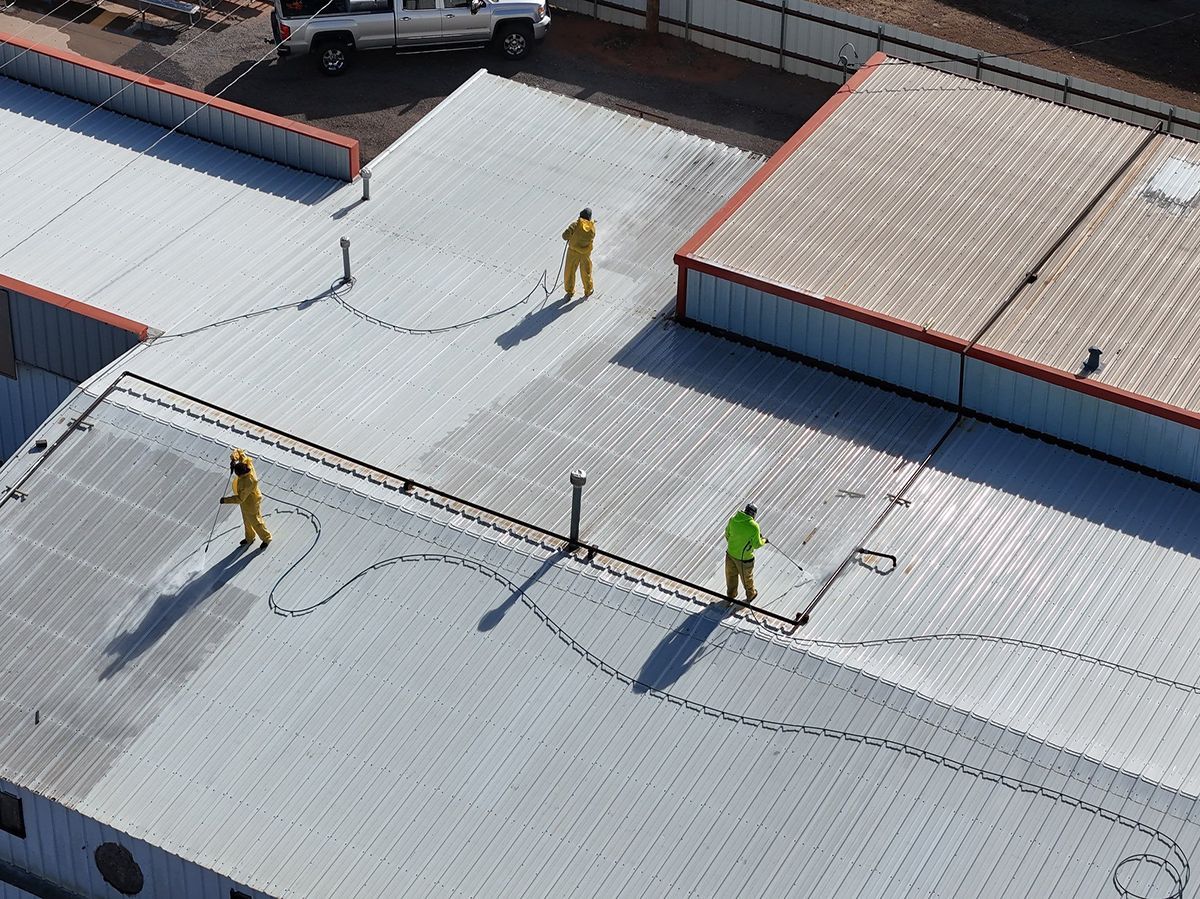 Three workers in yellow suits spraying a large, silver industrial roof; high angle view.
