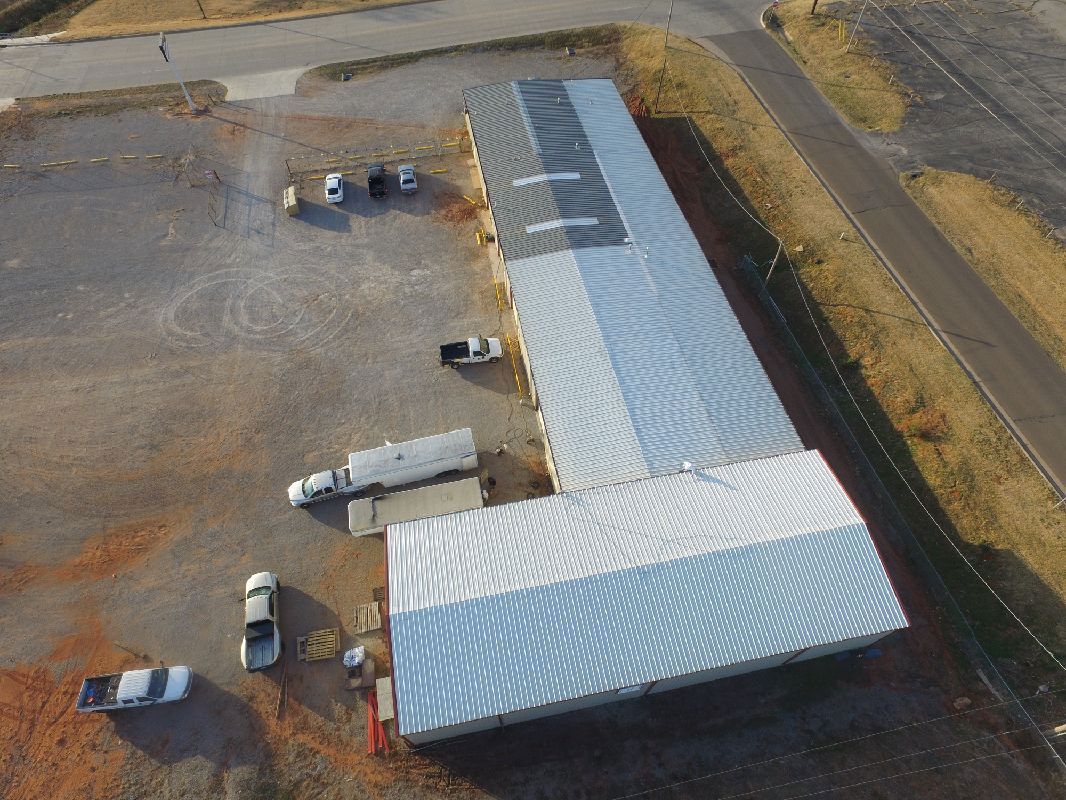 Overhead view of a large warehouse-style building with trucks and cars parked nearby on a dirt lot.