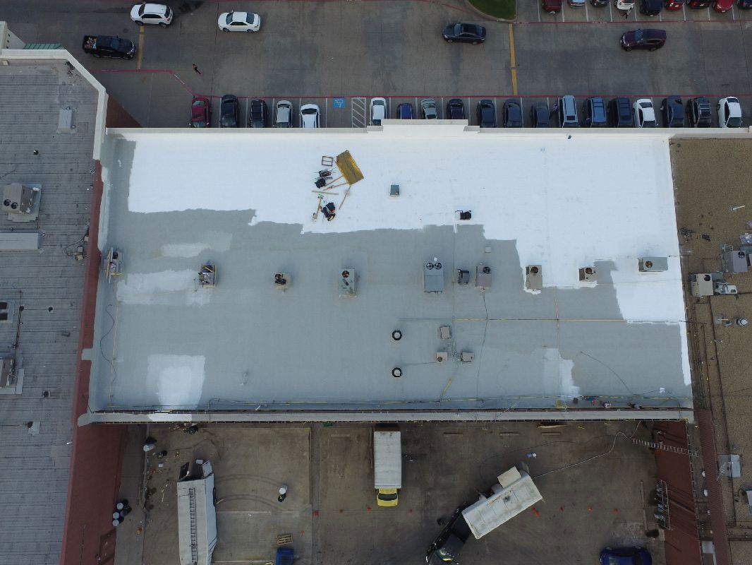 Aerial view of a building roof, partially painted white and gray. Parked vehicles surround the building.