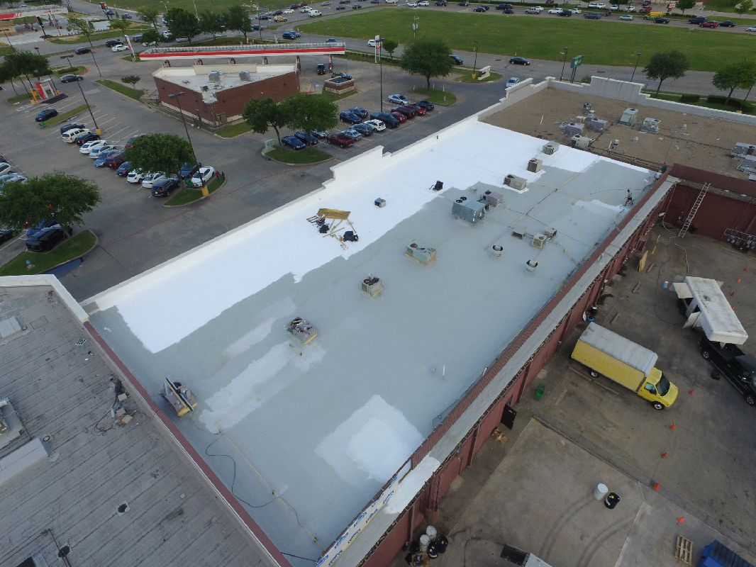 An aerial view of a commercial flat roof undergoing a white coating restoration, with parking and a yellow truck below.