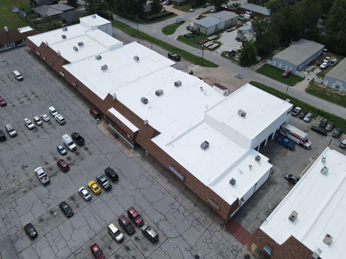 Aerial view of a long building with a white roof, surrounded by a parking lot with cars.