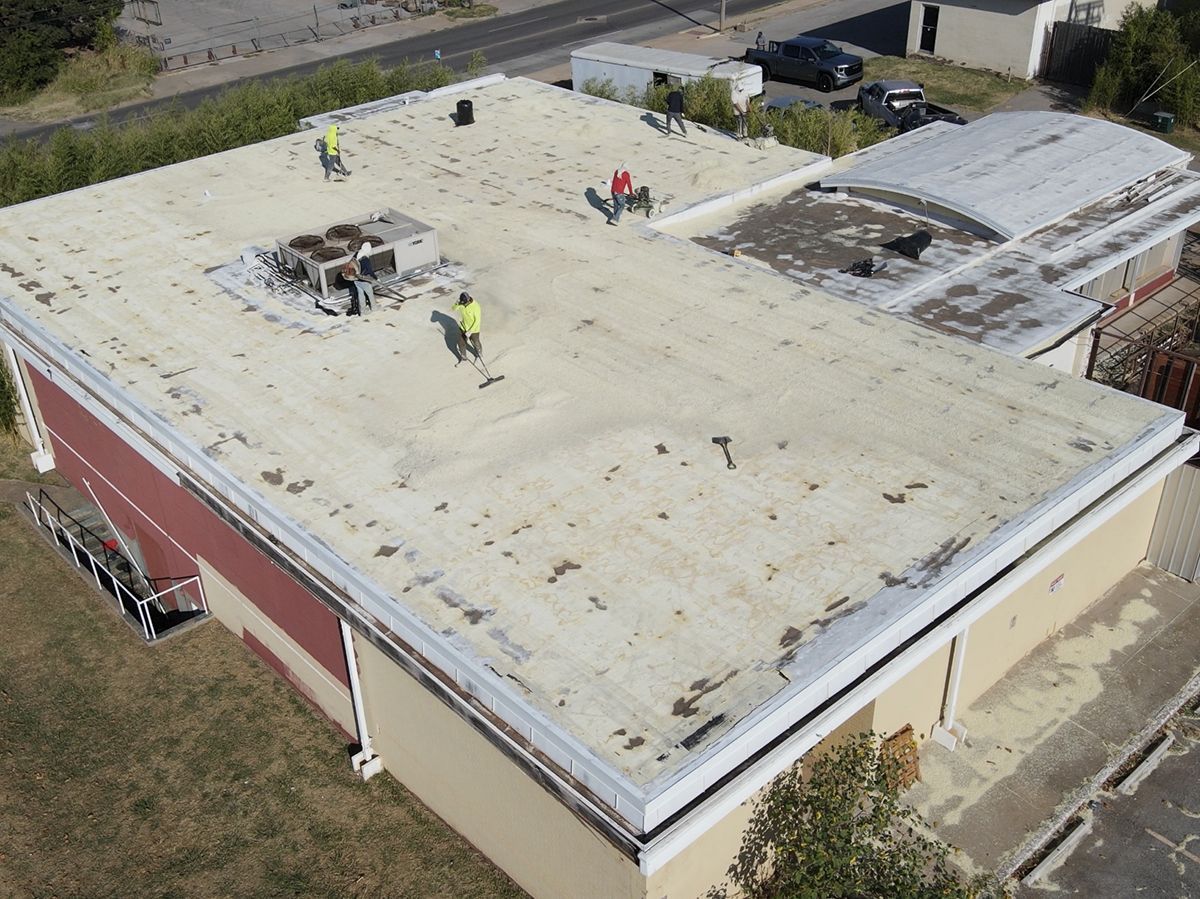 Workers on a flat commercial roof, repairing or inspecting. Beige roof, red and tan building. Sunny day.