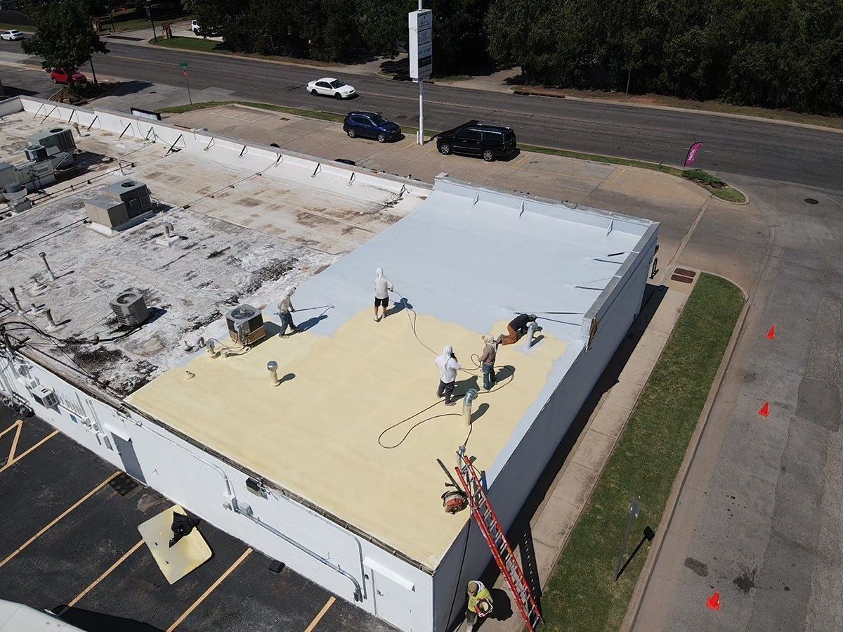 Workers on a flat commercial roof, applying a light-colored coating. Other sections are being prepared.