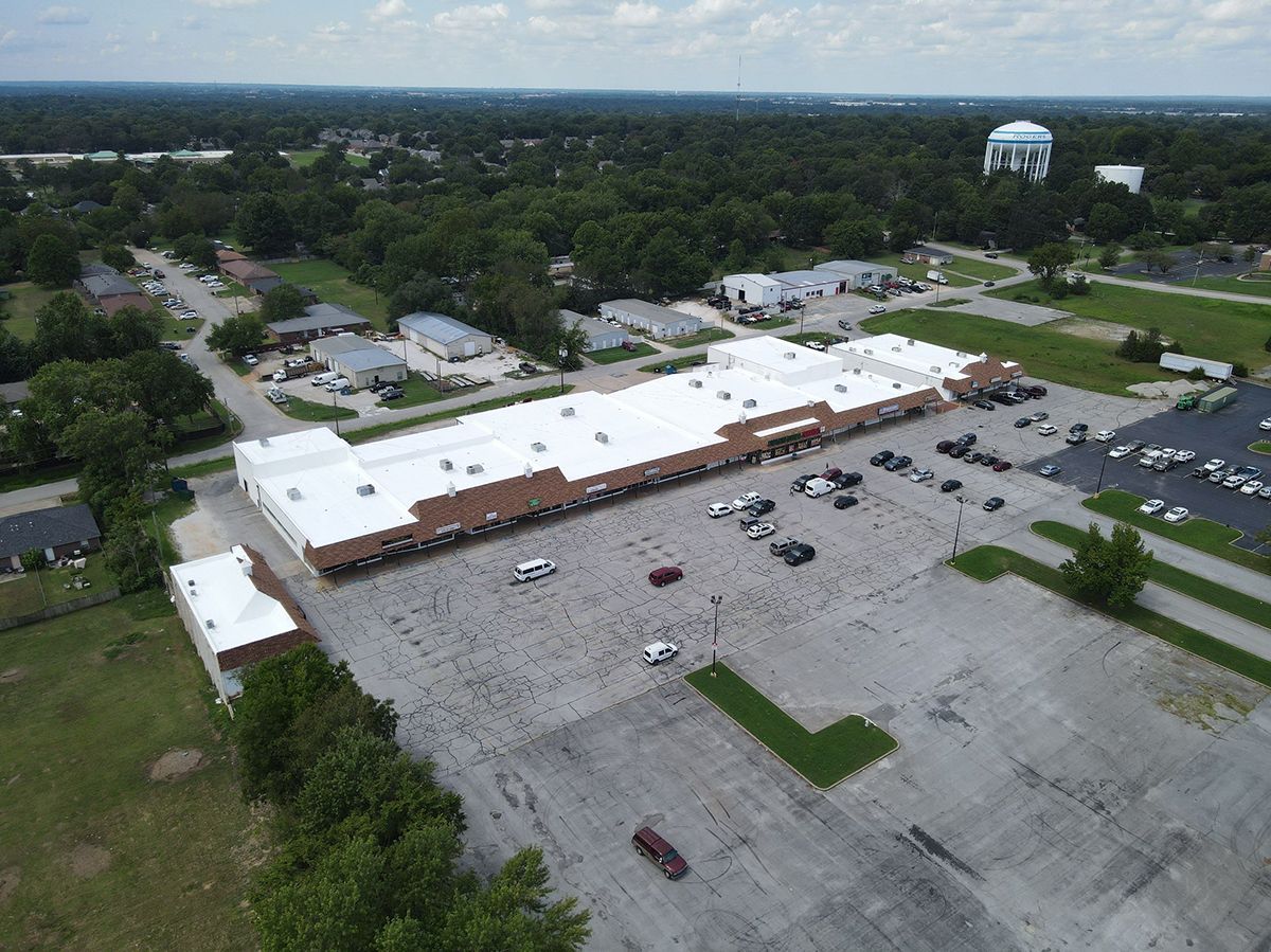 Aerial view of a long strip mall with a white roof and a large, mostly empty asphalt parking lot surrounded by trees.