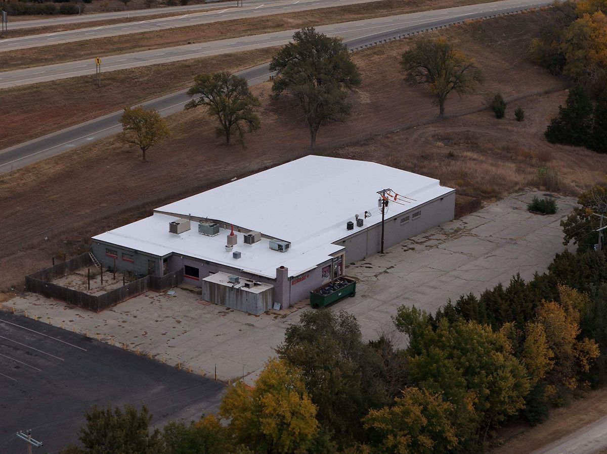 Aerial view of a tan industrial building with a white roof, near a road and some trees.