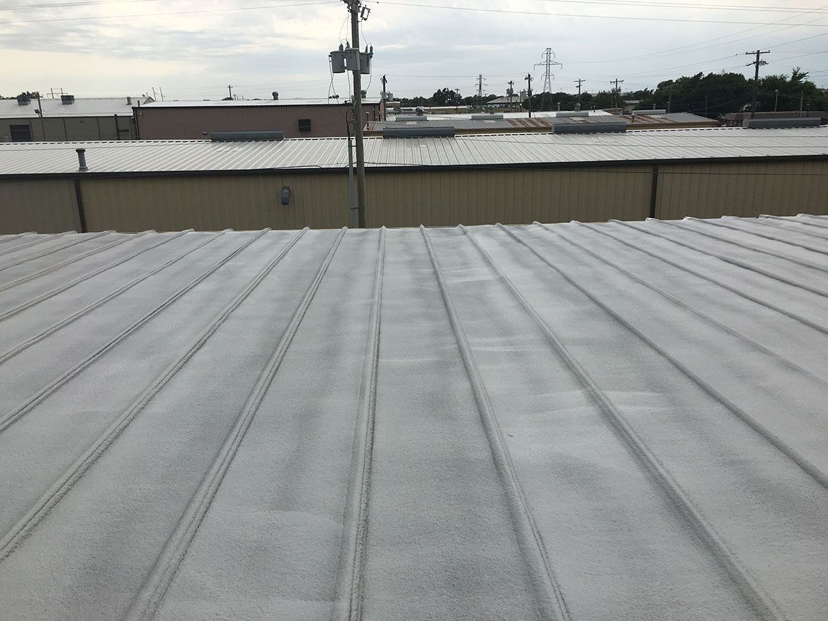 Metal roof with light-colored coating; view from above. Buildings and sky in background.