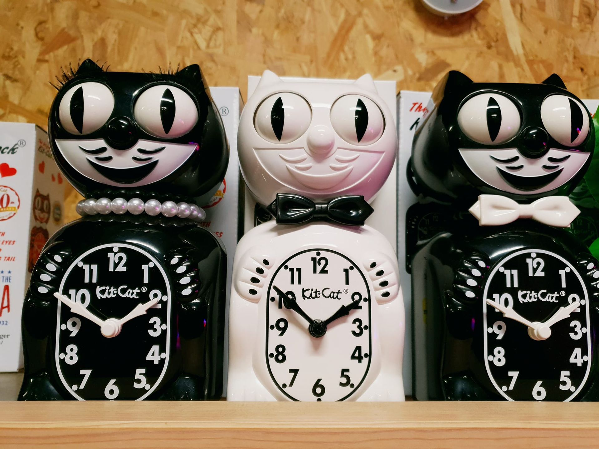 Trois horloges Kit-Cat Klock en forme de chat : deux noires avec des perles/nœuds papillon, une blanche avec un nœud papillon.