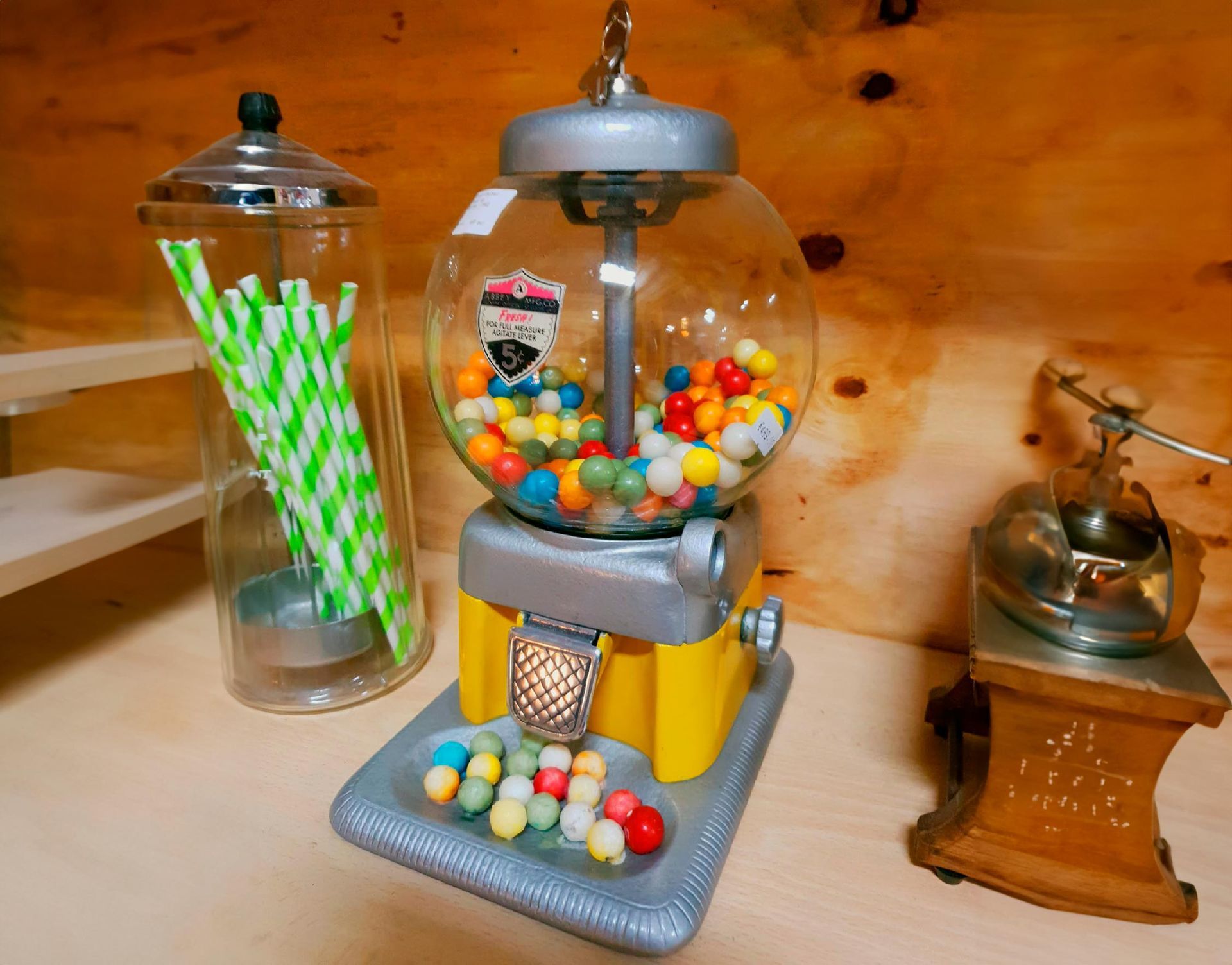 Distributeur de bonbons à mâcher avec des bonbons assortis, à côté d'un bocal en verre contenant des pailles et d'un petit broyeur en métal, sur une surface en bois.