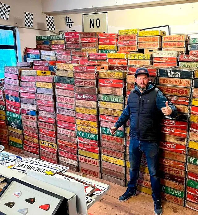 Un homme pose, pouces levés, devant un mur de caisses vintage colorées de Pepsi et de Coca-Cola.