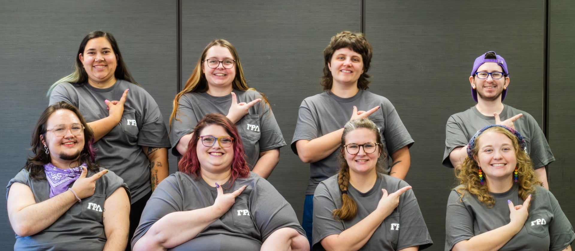 Eight people in matching gray shirts pose smiling and holding up the GRL hand sign against a dark wall.
