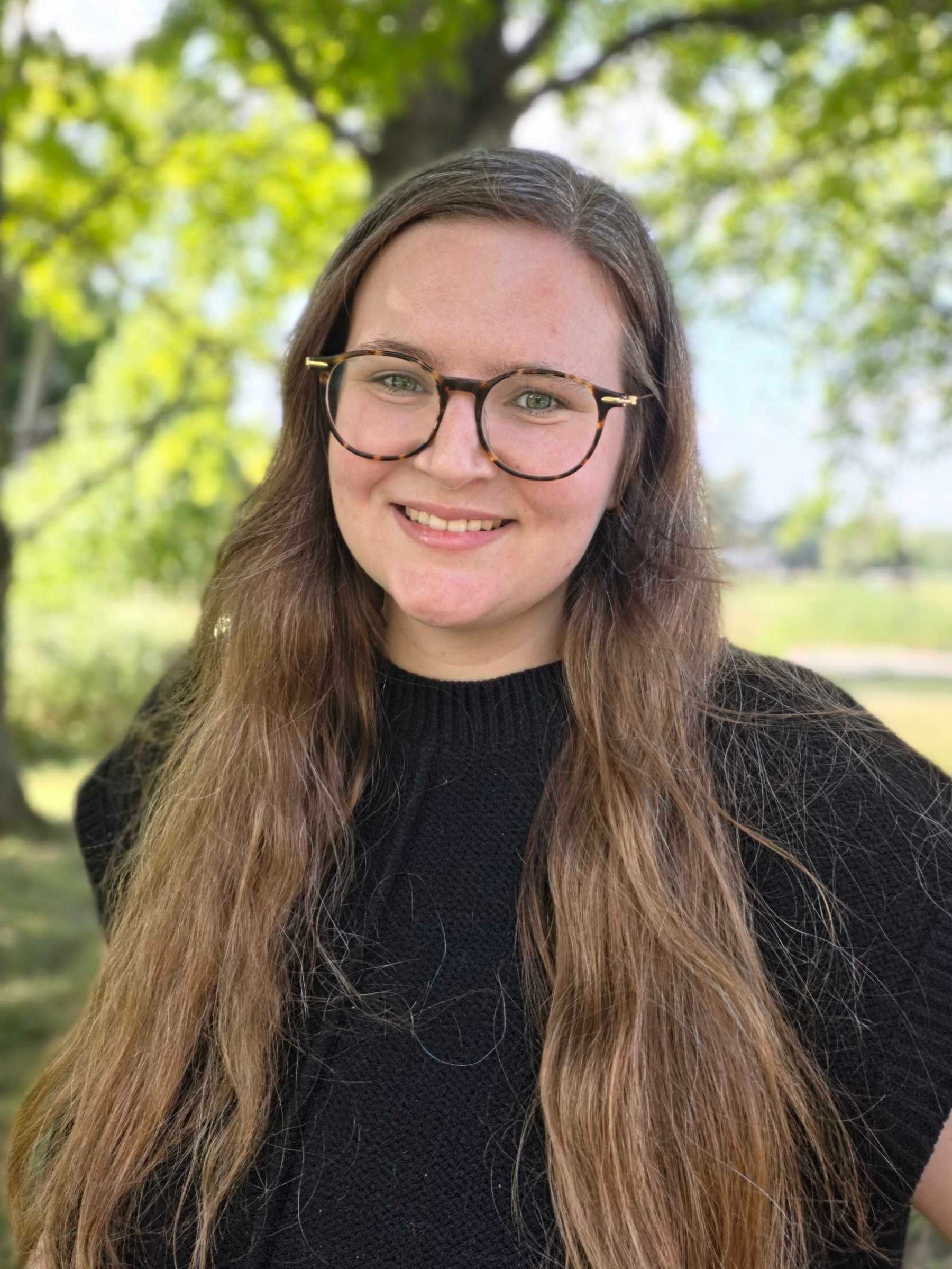 Smiling person with glasses and long brown hair outdoors, wearing a black top with green trees behind.