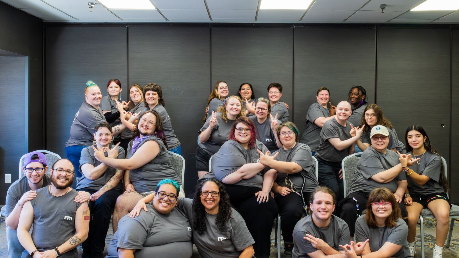 Group of smiling people posing together in a conference room with dark walls and bright overhead lights. They are leaning together and many are doing the linked GRL hand signal.