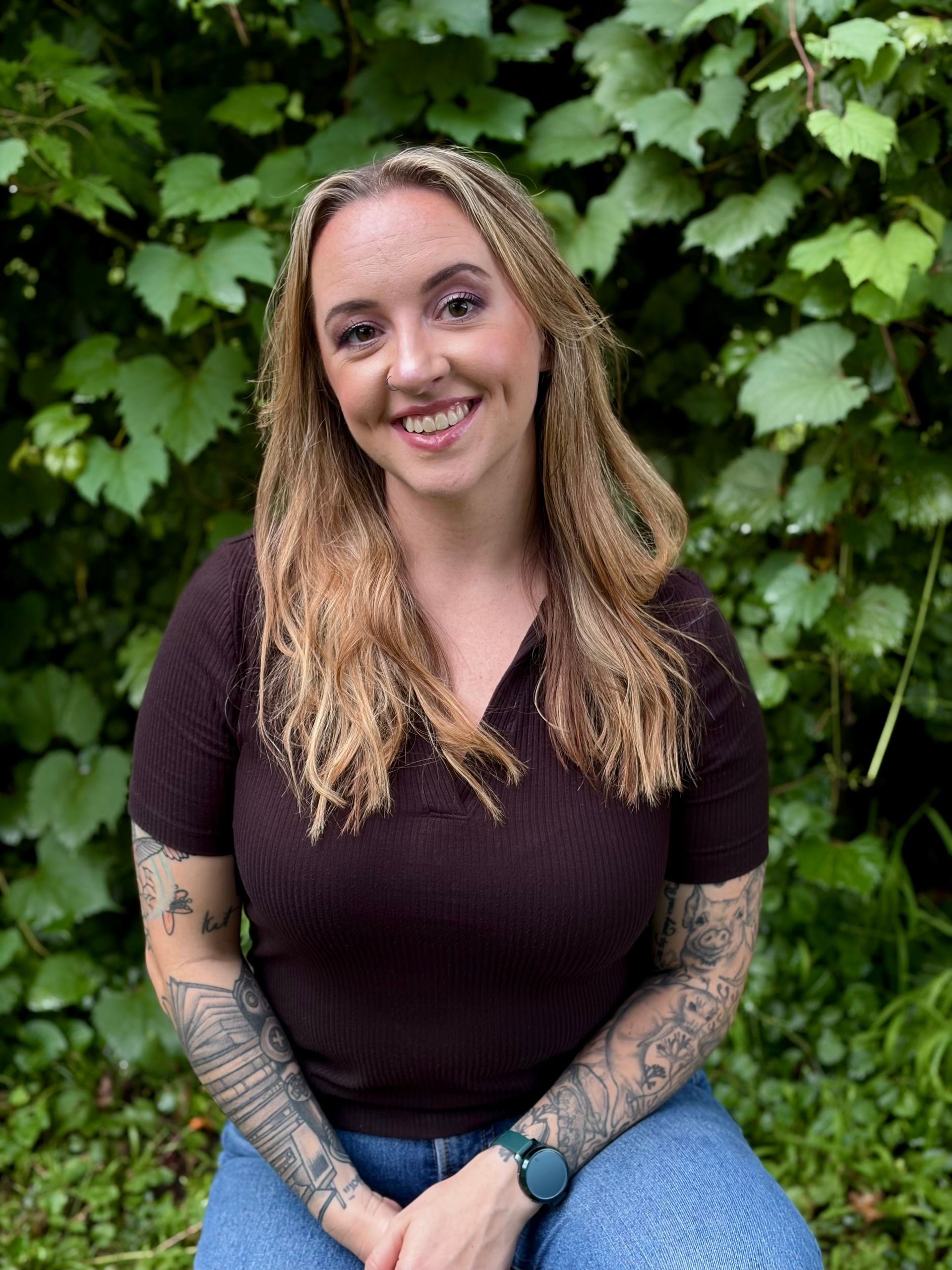 Smiling woman with tattooed arms sitting outdoors in front of green foliage, wearing a dark top and blue jeans