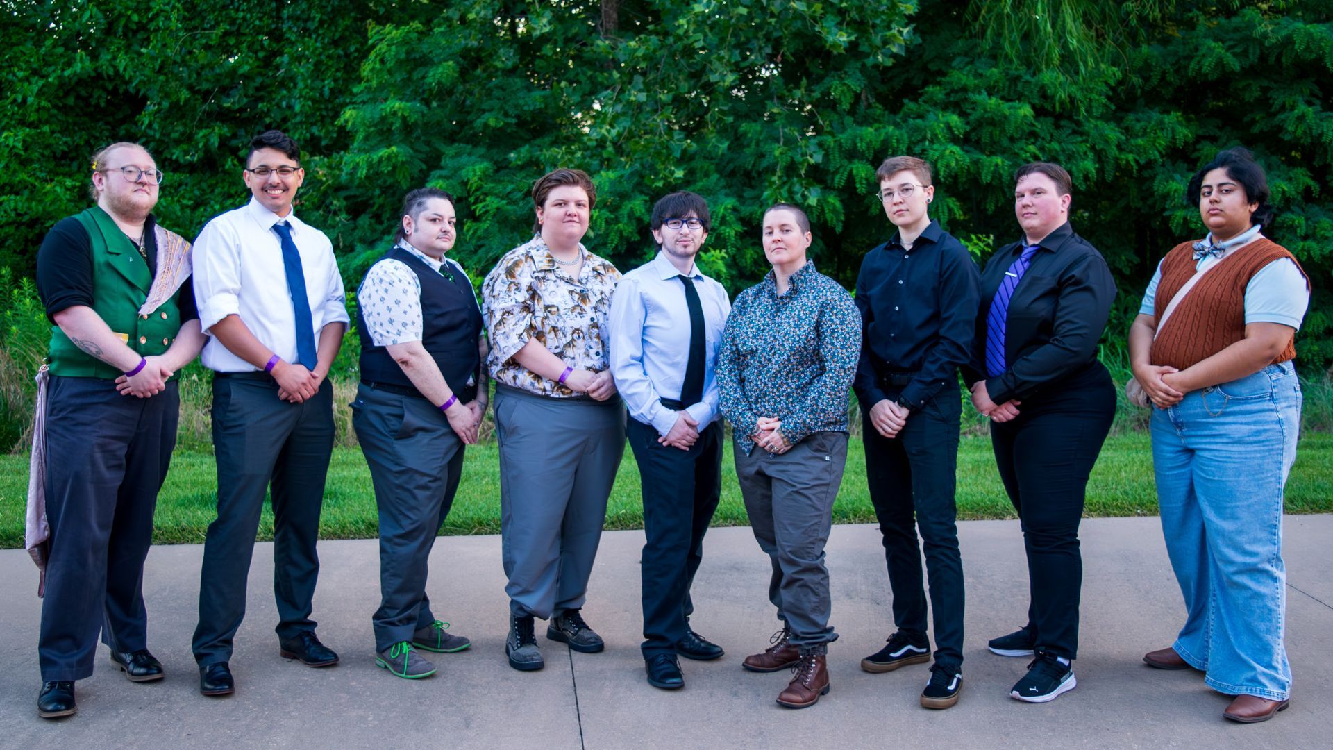 Nine people standing in a row outdoors on a sidewalk, dressed in semi-formal outfits.