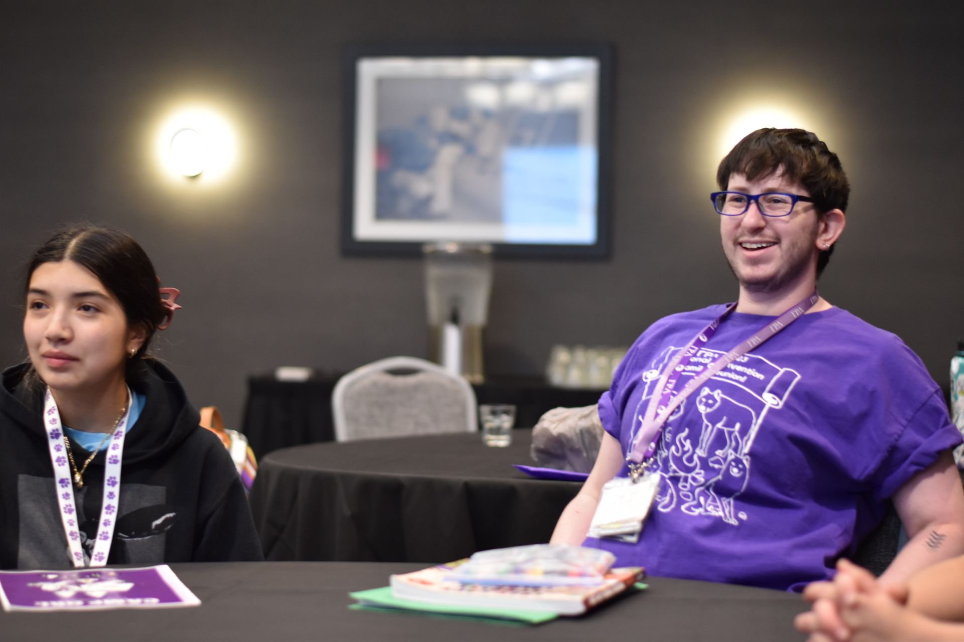 A dark haired person in a purple shirt seated at a table in a conference room. He is smiling with a lanyard around his neck.