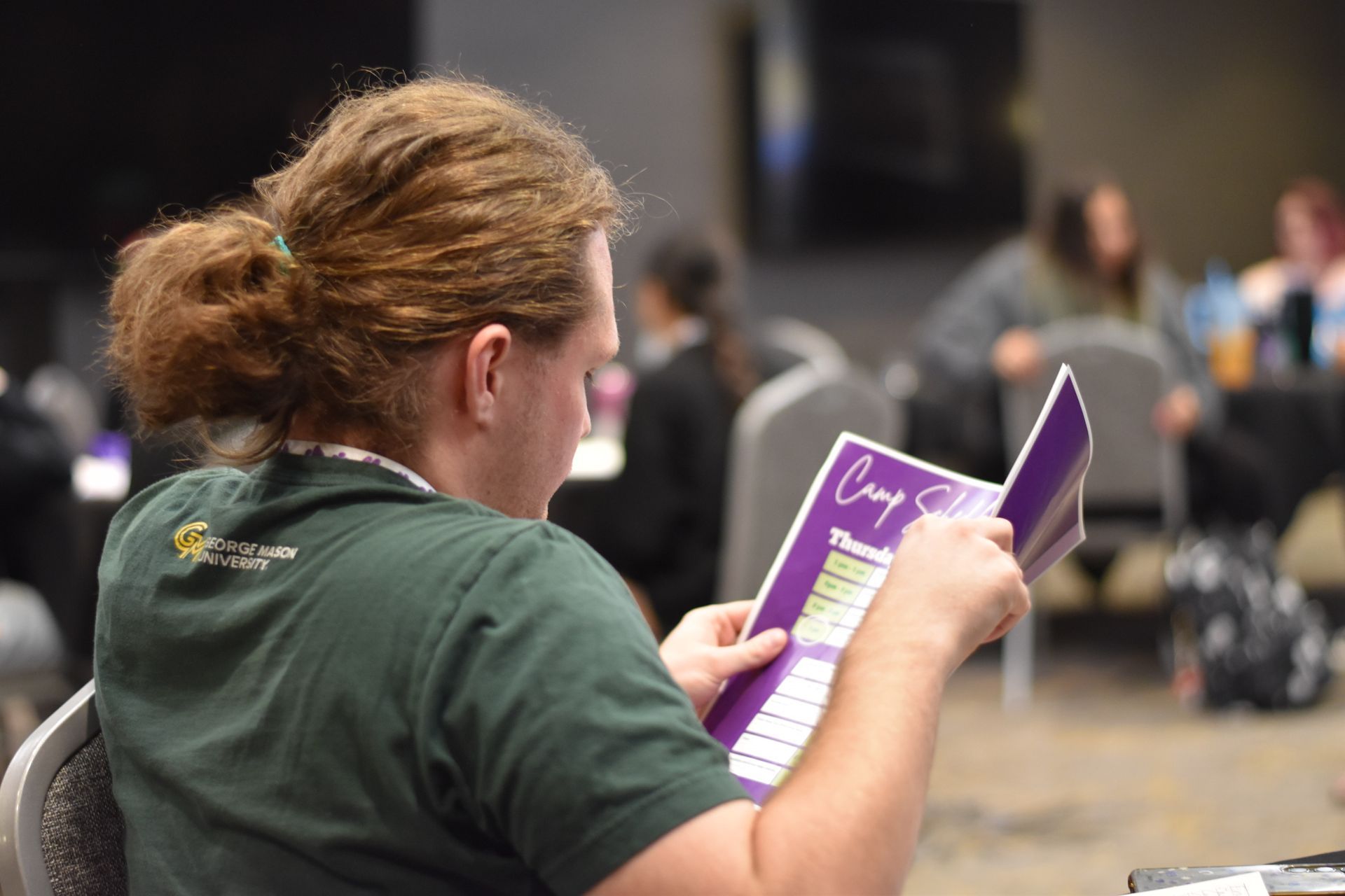 Person reading a purple booklet in a conference room, seated with people blurred in the background