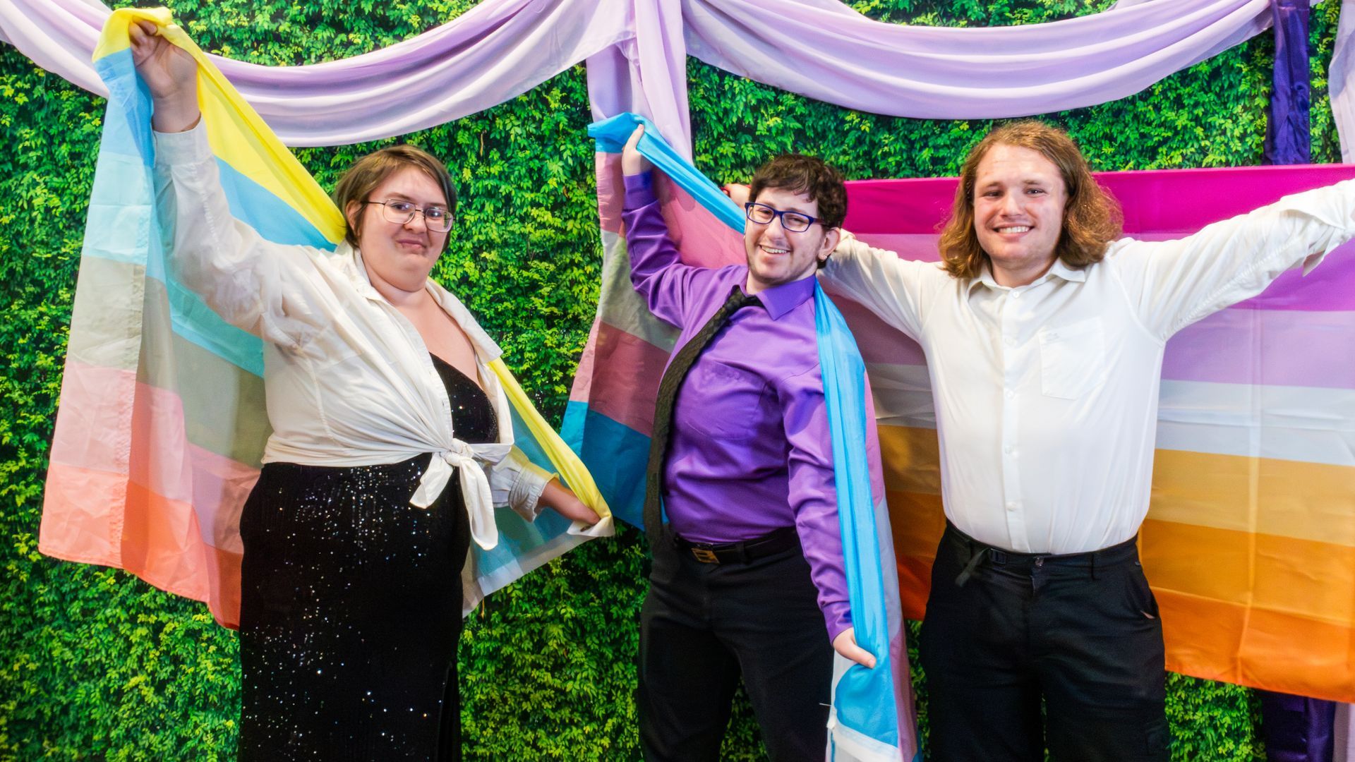 Three people smiling and holding the genderflux, trans, and lesbian pride flags in front of purple drapes and greenery