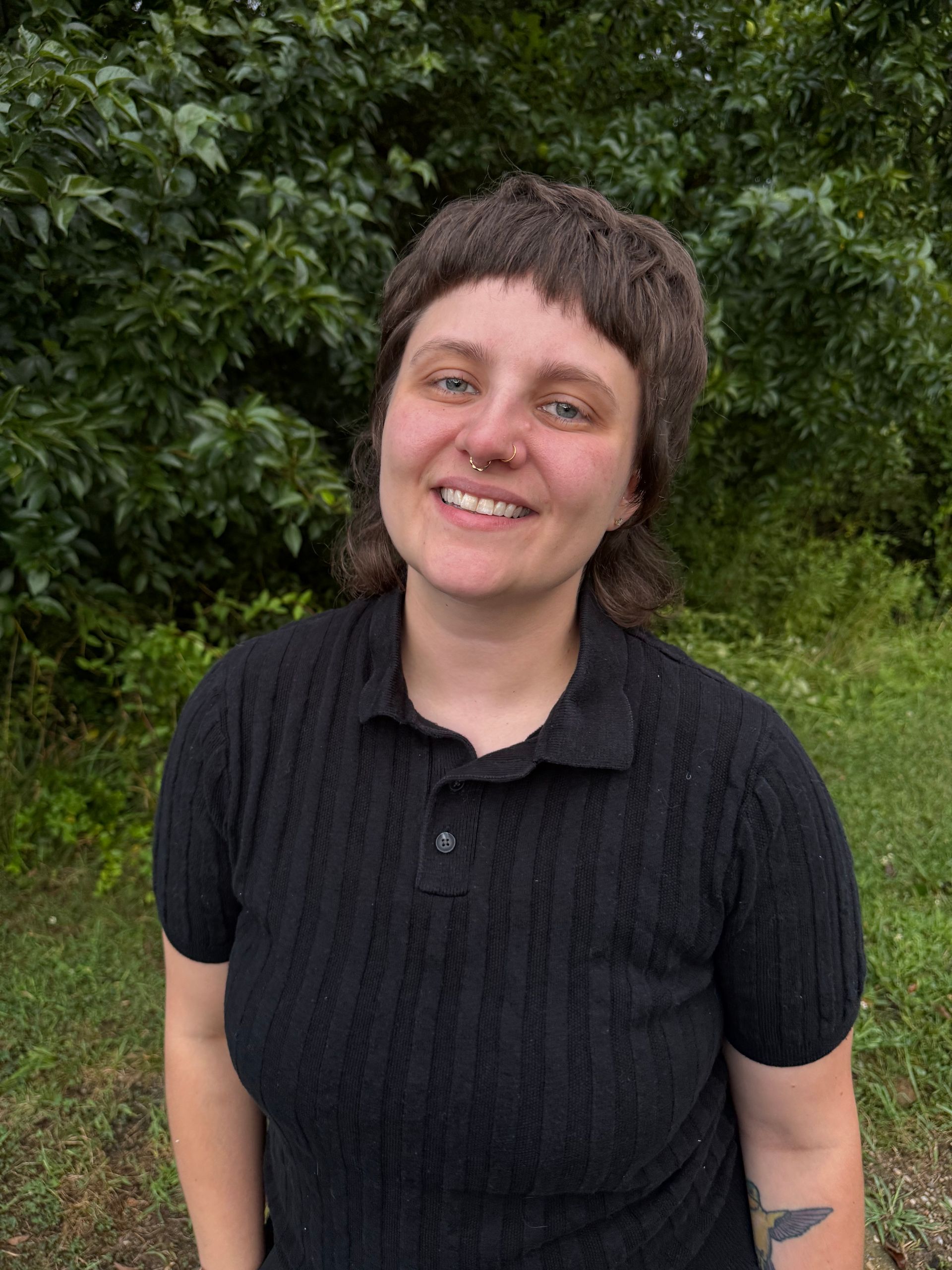 Smiling person in a black polo shirt standing outdoors in front of green foliage