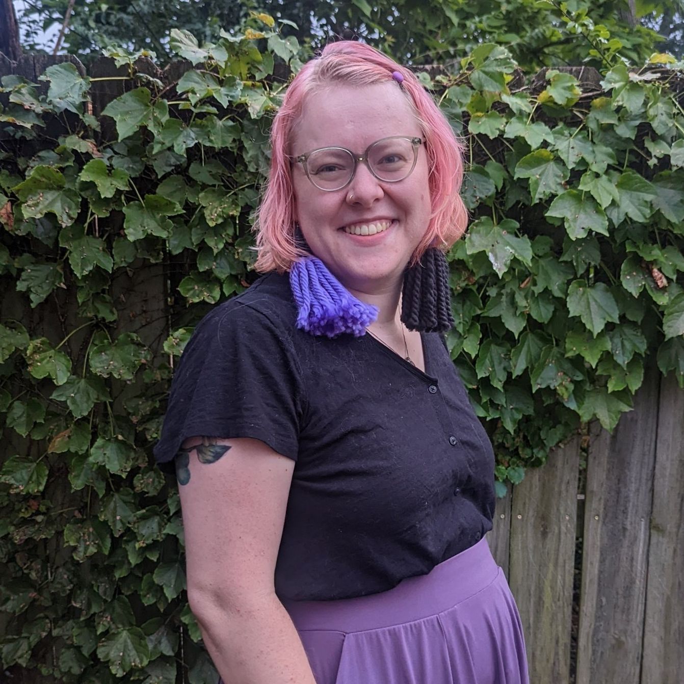 Person with pink hair and glasses smiles in front of ivy-covered fence, wearing a black top and purple skirt and purple and black earrings.