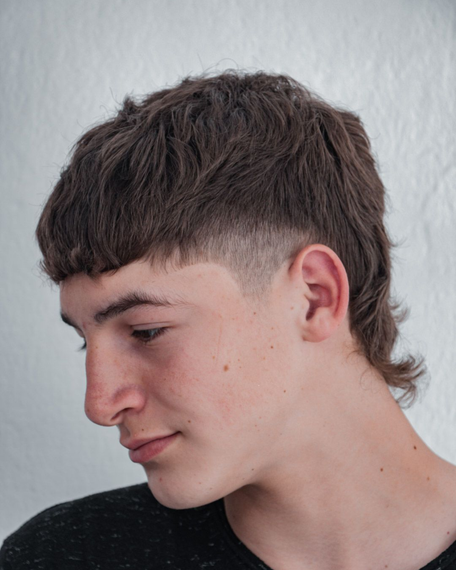 Perfil de una persona joven con cabello corto y oscuro sobre un fondo gris claro mostrando un corte mullet