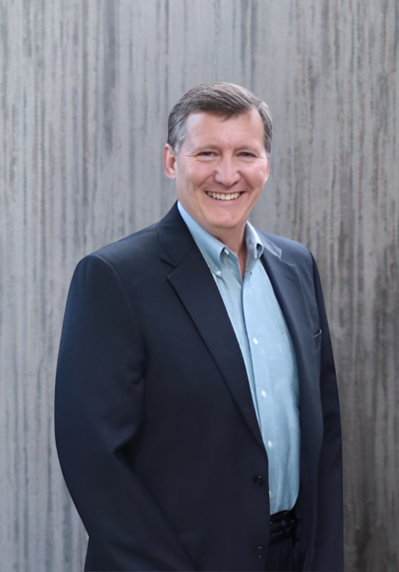 Man in a suit smiles, standing outside against a gray wall.