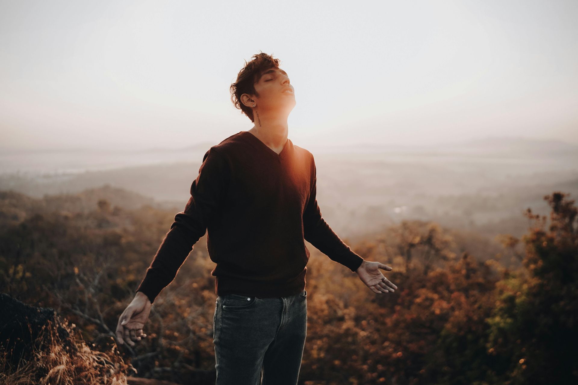 Person standing outdoors at sunrise with arms open, facing the light over a hazy landscape