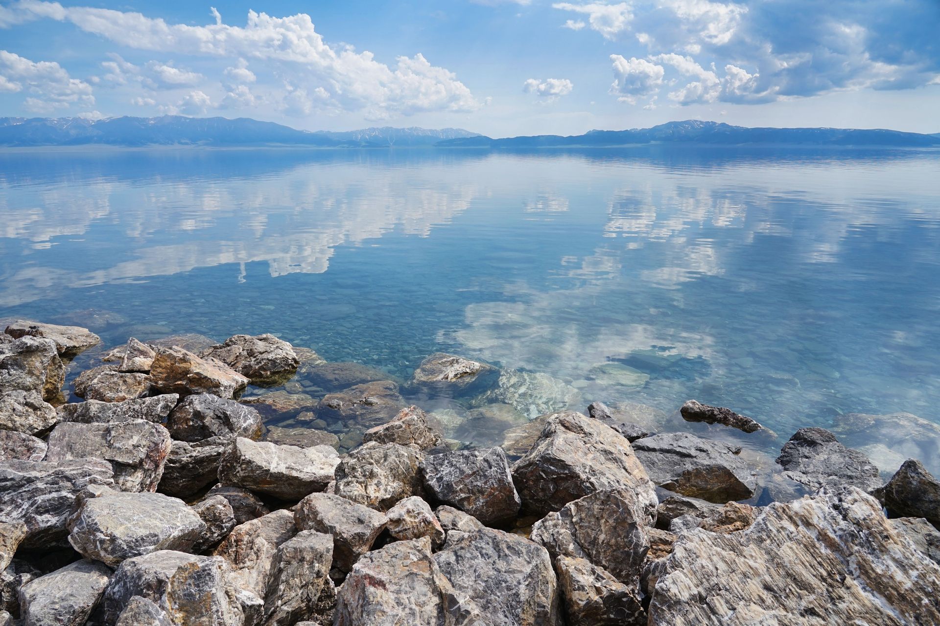 Rocky shoreline beside a calm blue lake with cloudy sky and distant mountains reflected in the water