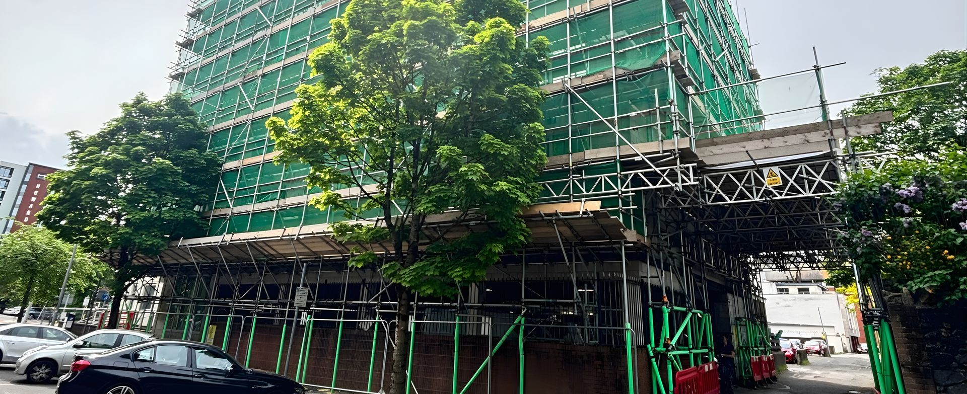 Scaffolding around a green building beside a tree-lined street with parked cars