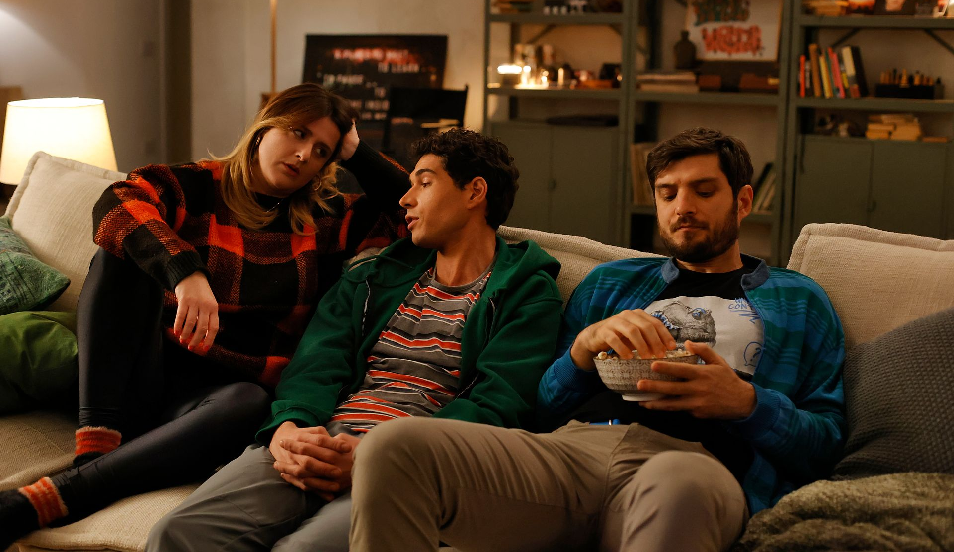 Drei Freunde sitzen auf einem Sofa und schauen sich etwas an. Einer isst Popcorn, einer lehnt sich an und der andere schaut zum Zuschauer.