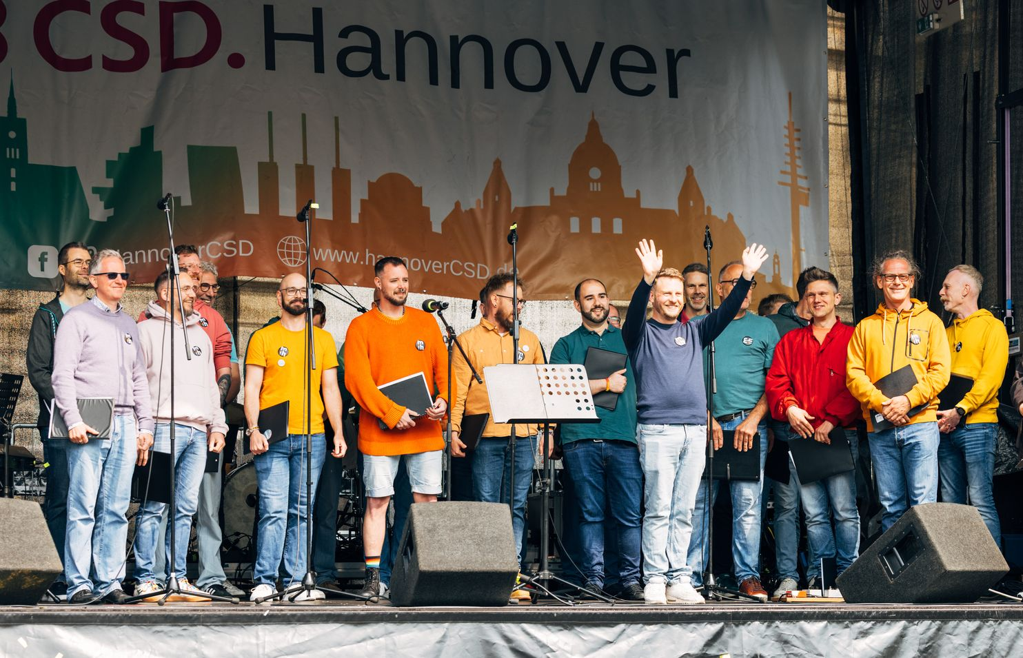 Eine Gruppe von Männern winkt auf der Bühne bei einer CSD-Veranstaltung in Hannover vor der Skyline der Stadt im Hintergrund.