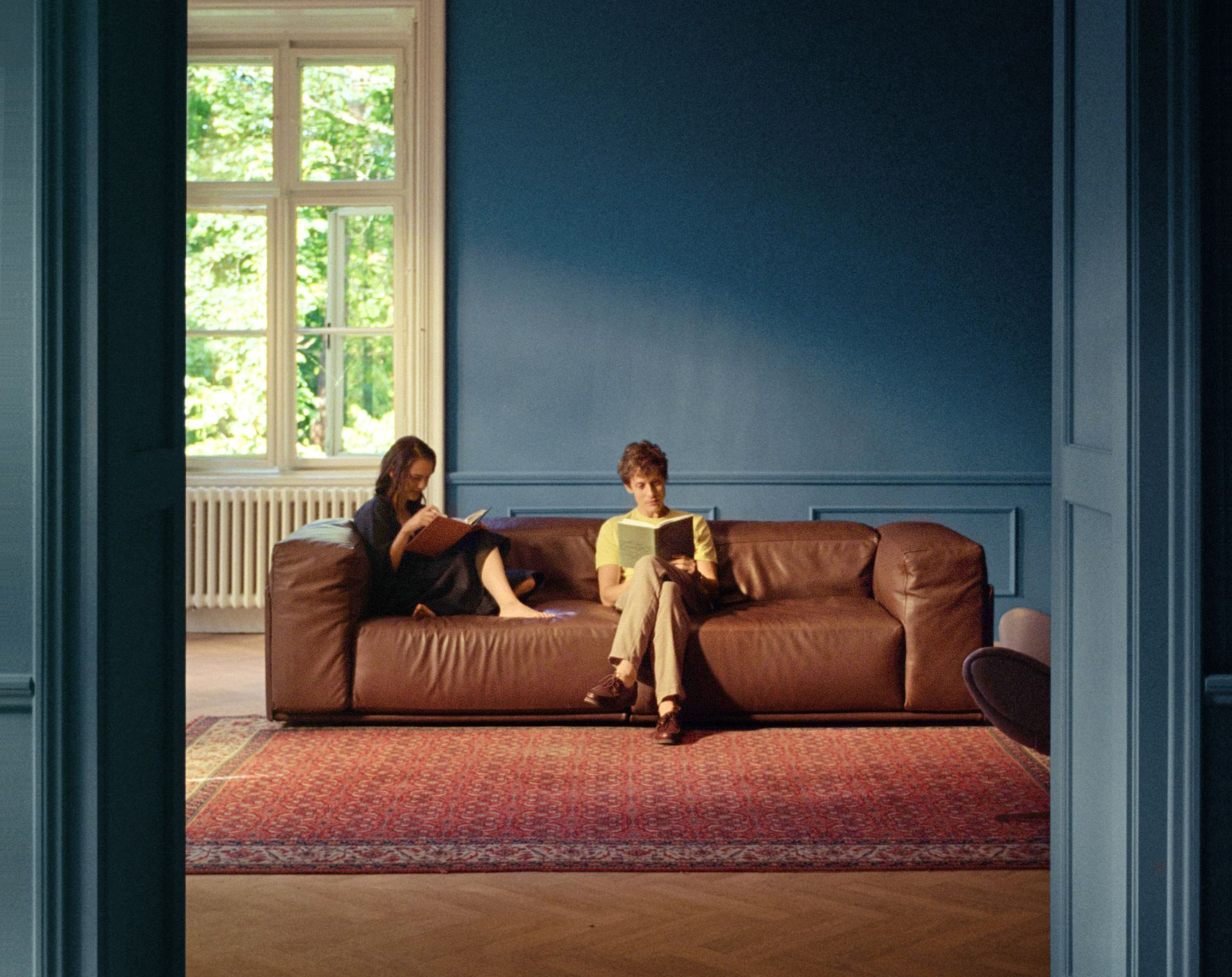 Zwei Personen lesen auf einem braunen Ledersofa in einem Raum mit rotem Teppich und blauer Wand.
