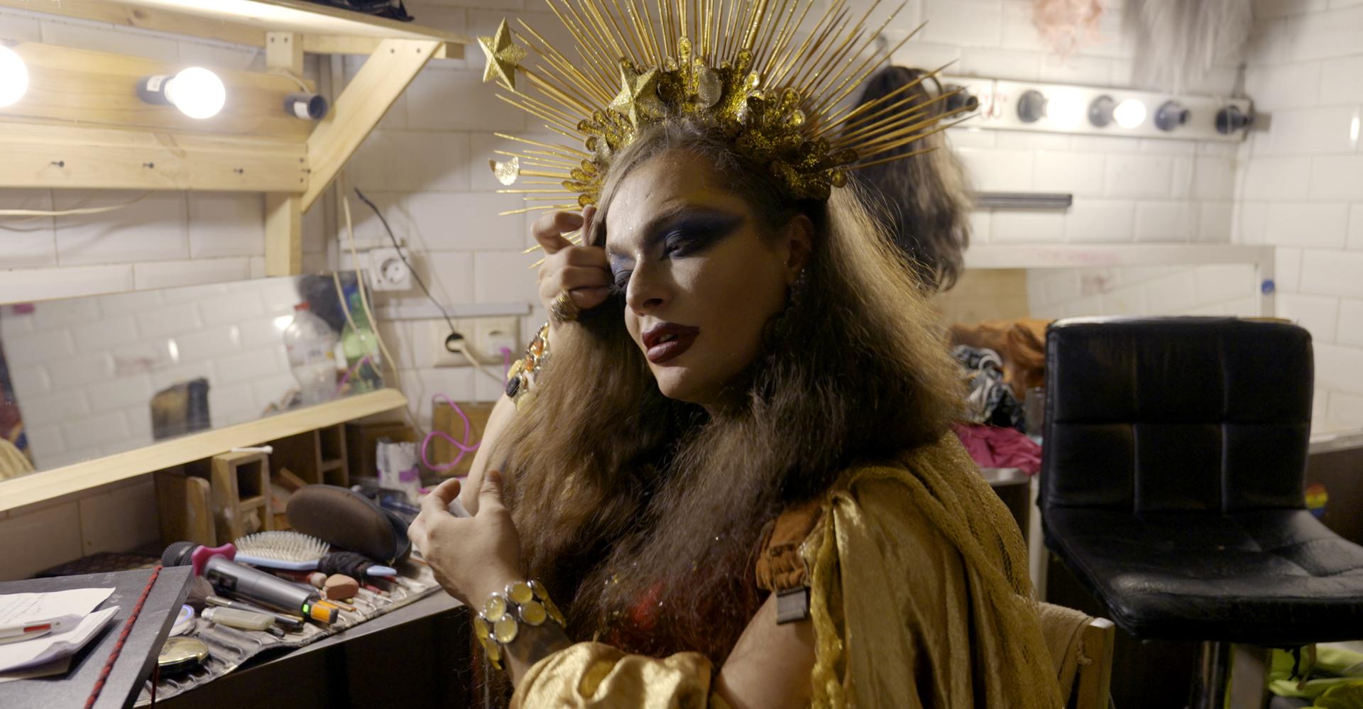 Eine Person in aufwendiger goldener Kleidung und Make-up rückt in einer Umkleidekabine einen Kopfschmuck zurecht.