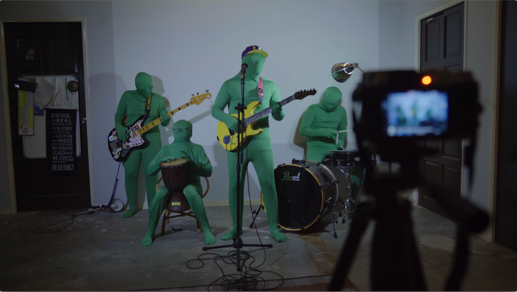 Eine Band in grünen Ganzkörperanzügen tritt in einem Raum auf und filmt mit einer Kamera auf einem Stativ.