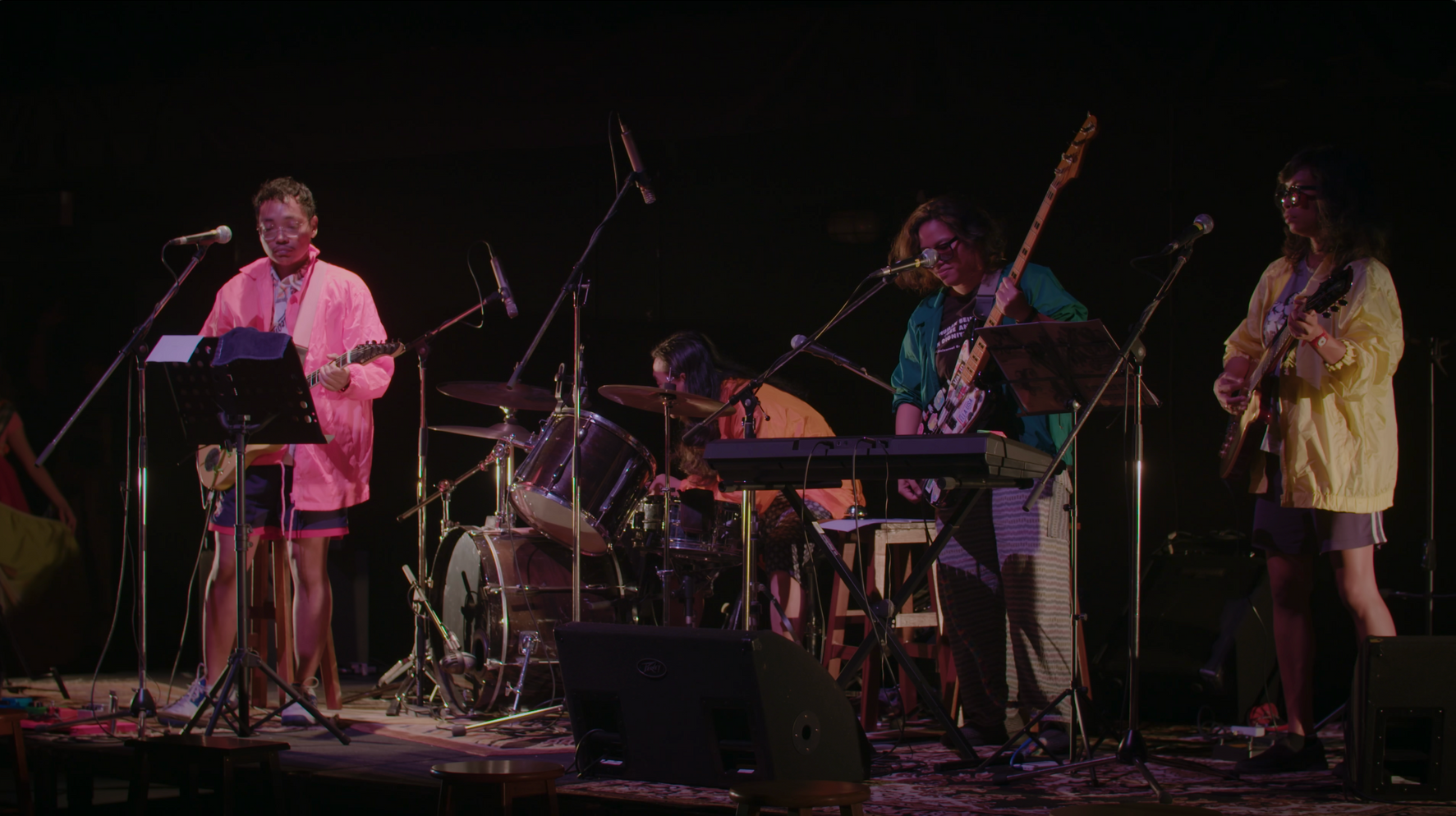 Band auf der Bühne bei dunkler Beleuchtung, spielt Instrumente. Die Musiker tragen bunte Kleidung in Rosa, Orange, Grün und Gelb.