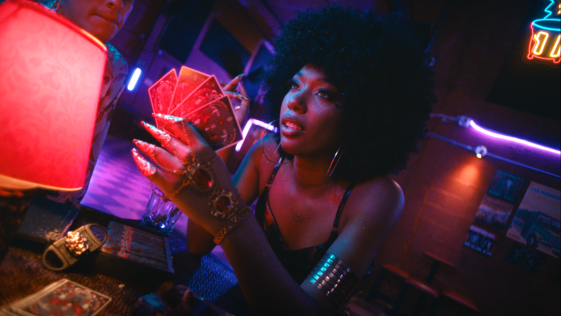 Eine Person mit Afrofrisur mischt Karten in neonbeleuchteter Bar, trägt ein Armband und hat lange, manikürte Nägel.