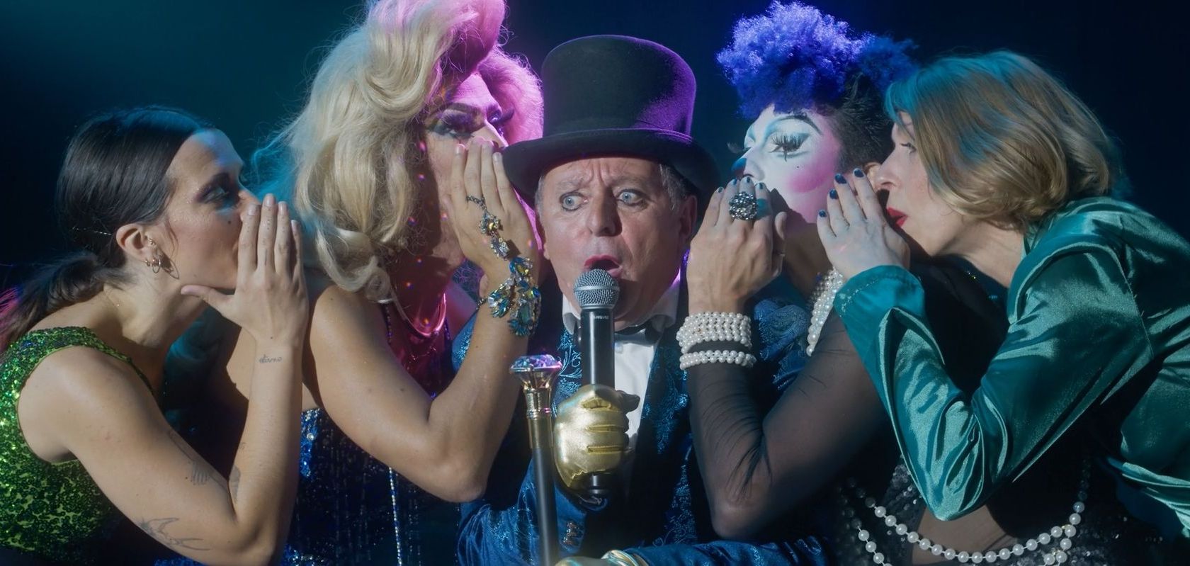 Eine Gruppe Drag Queens flüstert in das Mikrofon eines Mannes.