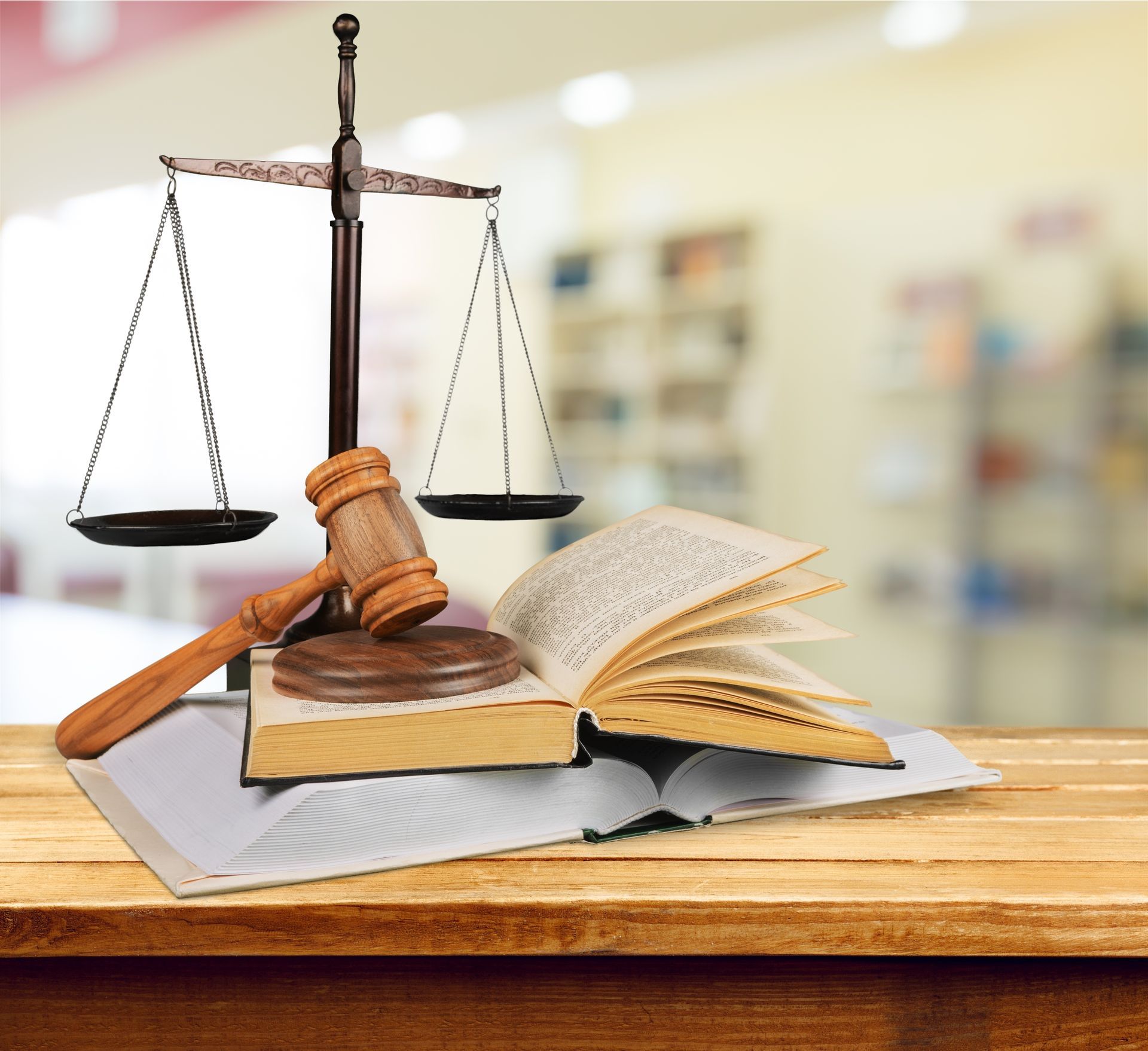 Scales of justice, gavel, and open law books on a wooden table, blurry background of a library.