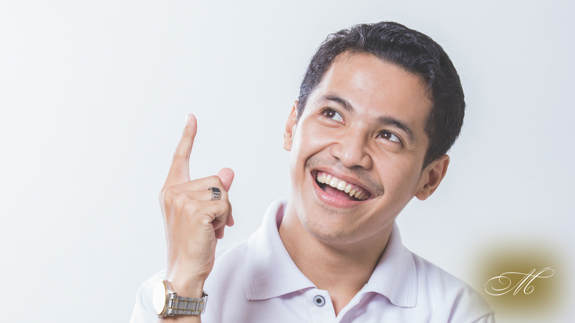 Man with dark hair smiles, pointing upwards; wearing a white polo shirt and watch.