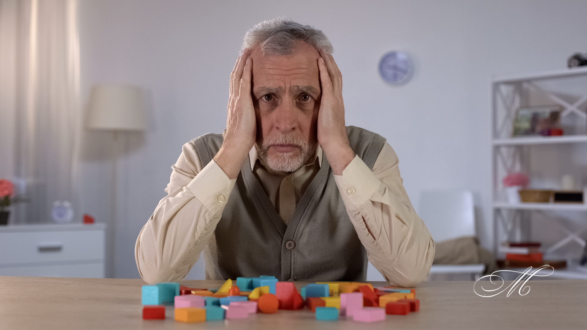 An elderly man is sitting at a table with his hands on his head.
